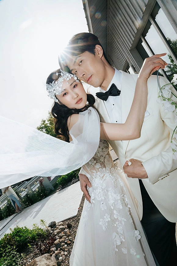 Couple portrait of bride and groom embrace, her veil blowing in sun flare beside a modern building walkway, holding a bouquet