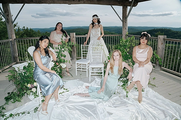 Bridal party portrait of bride with bridesmaids in mix-and-match satin dresses, posing on an outdoor deck with forested hills behind