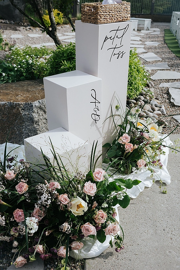 Petal toss station with petal toss sign in calligraphy on white pedestals, woven basket and rose florals along a garden path