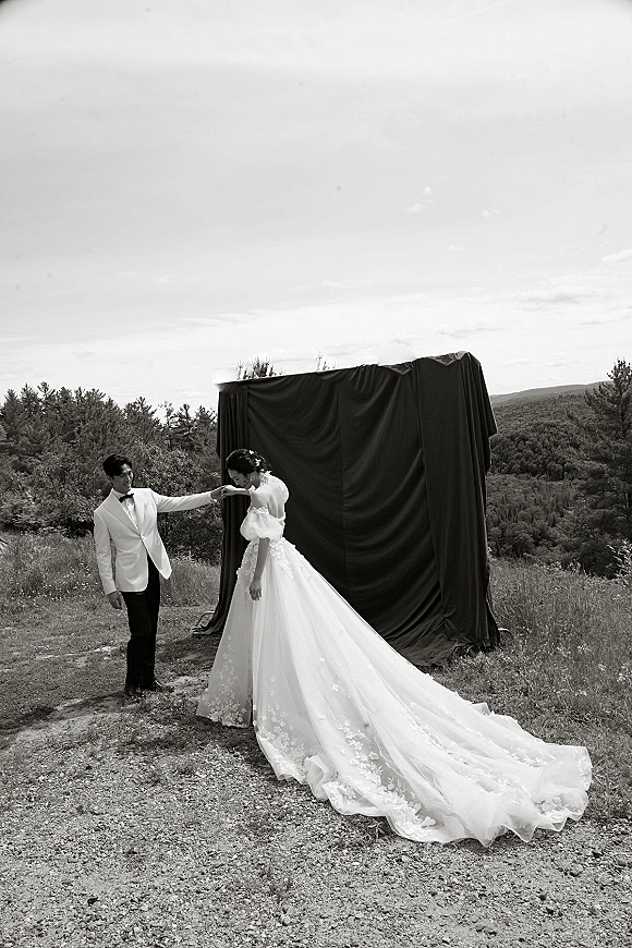 Couple portrait of bride and groom holding hands, her lace applique gown with long train and puff sleeves set against mountain meadow backdrop
