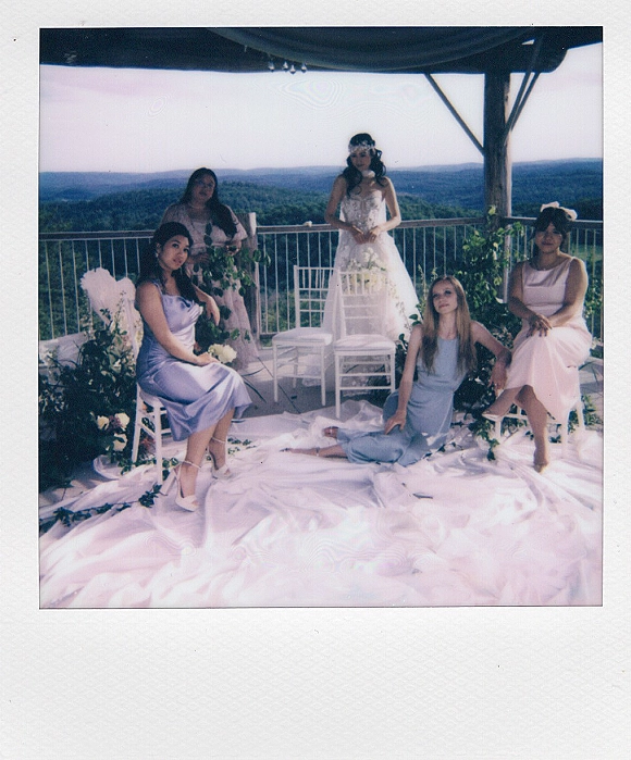 Bridal party portrait of bride with bridesmaids in pastel dresses, seated on an outdoor terrace with a mountain and forest view, bouquet accent