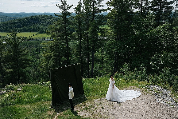 Couple portrait of bride with long train and groom in white dinner jacket posing by green fabric backdrop, with mountains and forest behind