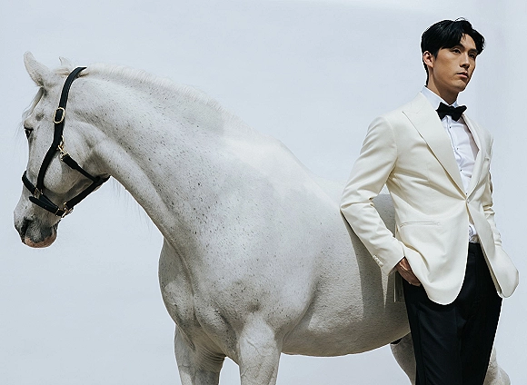 Groom portrait in a white tuxedo with black bow tie beside a white horse in halter, posed against an open sky backdrop