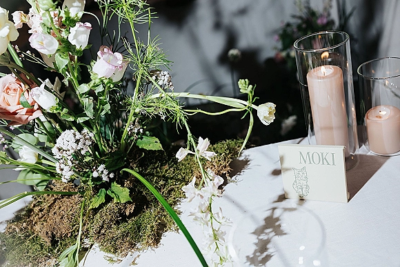 Wedding centerpiece with moss and roses, wildflowers, and greenery, accented by taper candles in glass cylinder vases on a white tablecloth