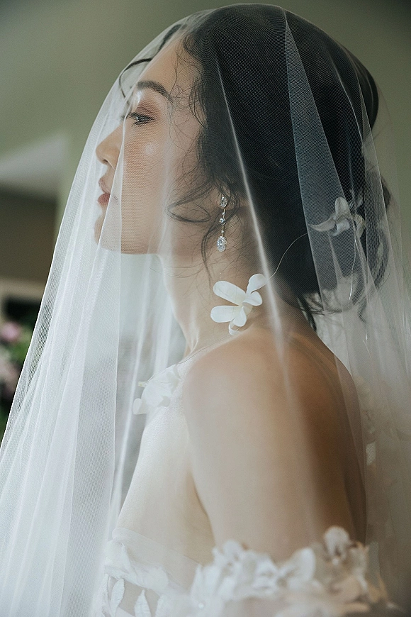 Bridal portrait of a bride in side profile with a veil over face, strapless dress, and drop earrings against a soft neutral wall