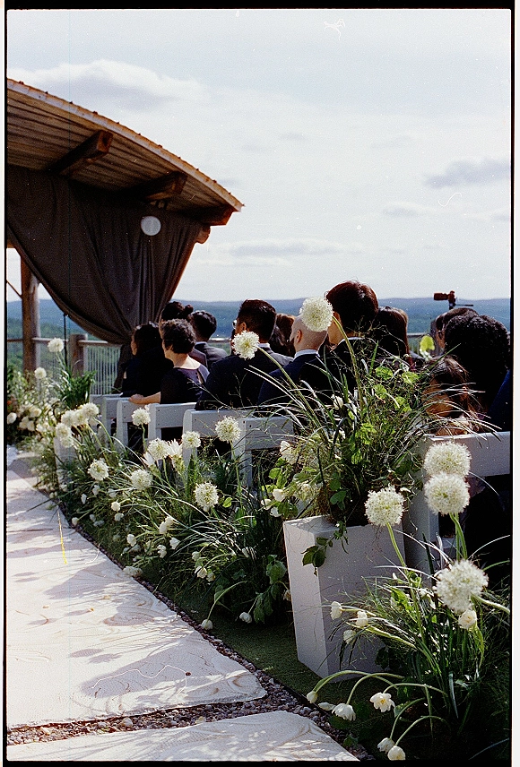 Outdoor ceremony setup with wedding aisle flowers and a white aisle runner leading to a draped wood pergola on a terrace with hills beyond