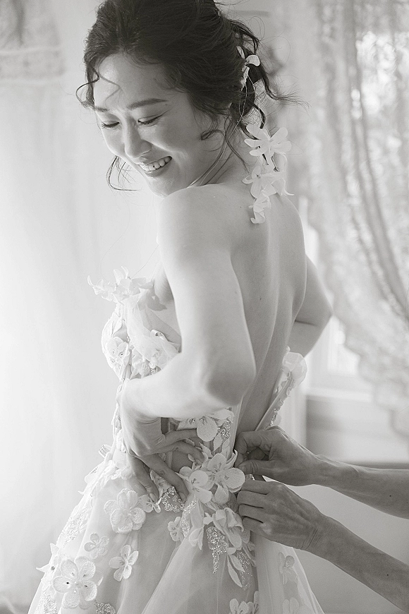 Bridal portrait of a bride in a low-back gown with floral appliqués and hair flowers, smiling in window light by sheer curtains indoors