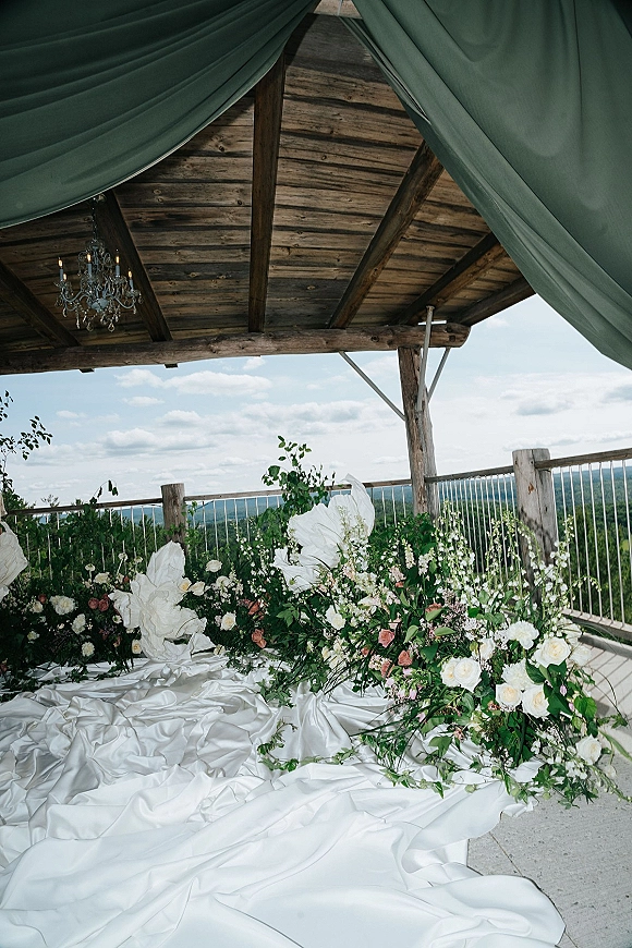 Wedding ceremony backdrop with white draped fabric, chandelier, and white rose greenery ground florals on a pergola patio with hills beyond