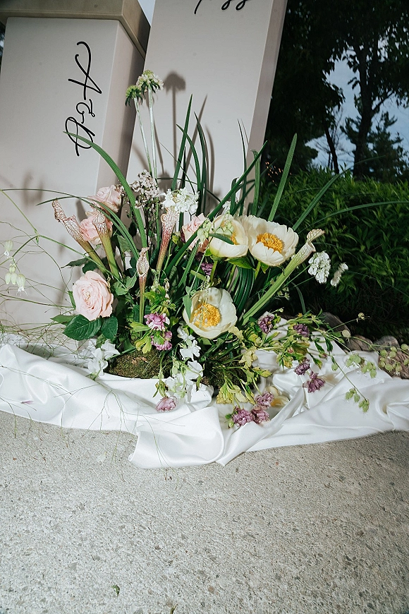 Welcome sign flowers on a wedding welcome sign with calligraphy lettering, roses and poppies in lush greenery, satin drape on concrete outdoors at night