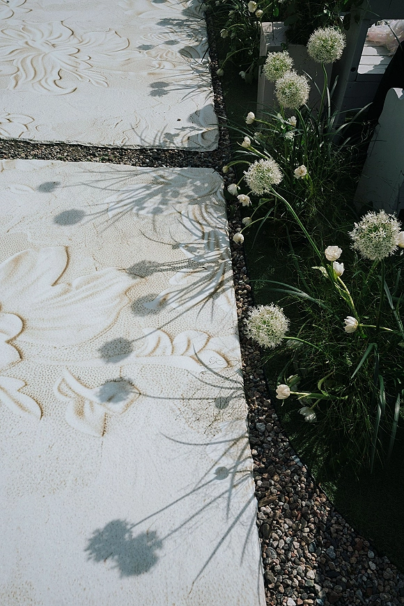 Ceremony aisle design with a wedding aisle runner on green grass, bordered by gravel and low allium and white tulip florals in sunlight