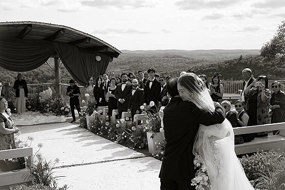 Wedding hug as bride in veil and gown embraces tuxedo groom on an outdoor terrace aisle, guests watching with mountains beyond