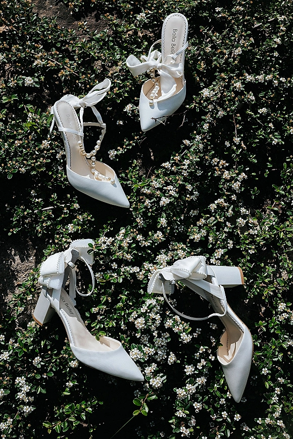 Bridal shoes in a wedding shoes flat lay, white heels with bow straps and pearl ankle straps on greenery with tiny flowers and sun shadows