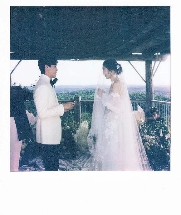 Wedding vows as bride in a strapless lace gown and veil faces groom in white tuxedo under a pavilion with mountain view and florals
