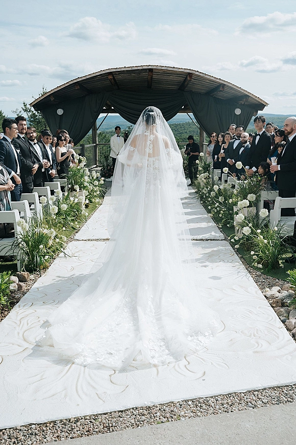 Wedding processional with bride walking down aisle in a cathedral veil, gown train trailing along a flower-lined runner toward a mountain-view arch