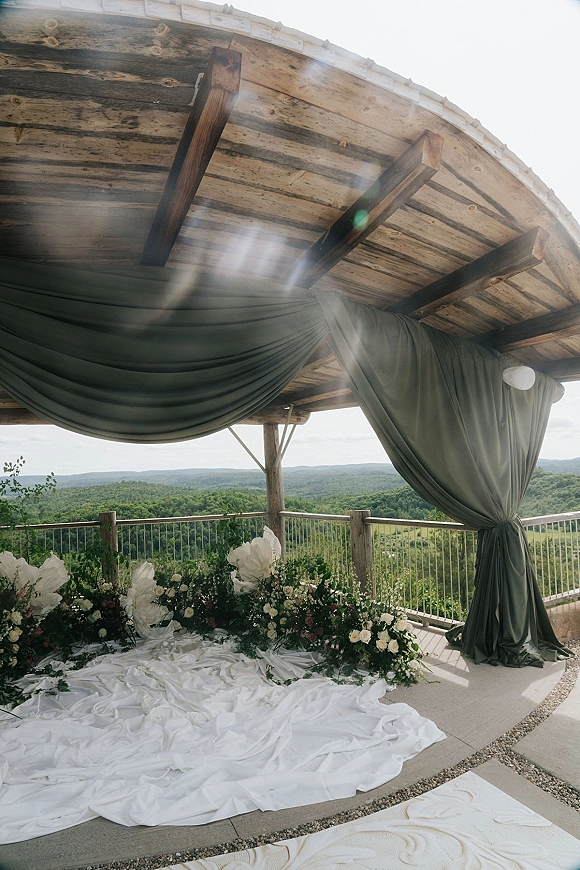 Ceremony backdrop with wedding ceremony draping, floral arrangements, and greenery on a wooden pavilion overlooking mountain hills and sky