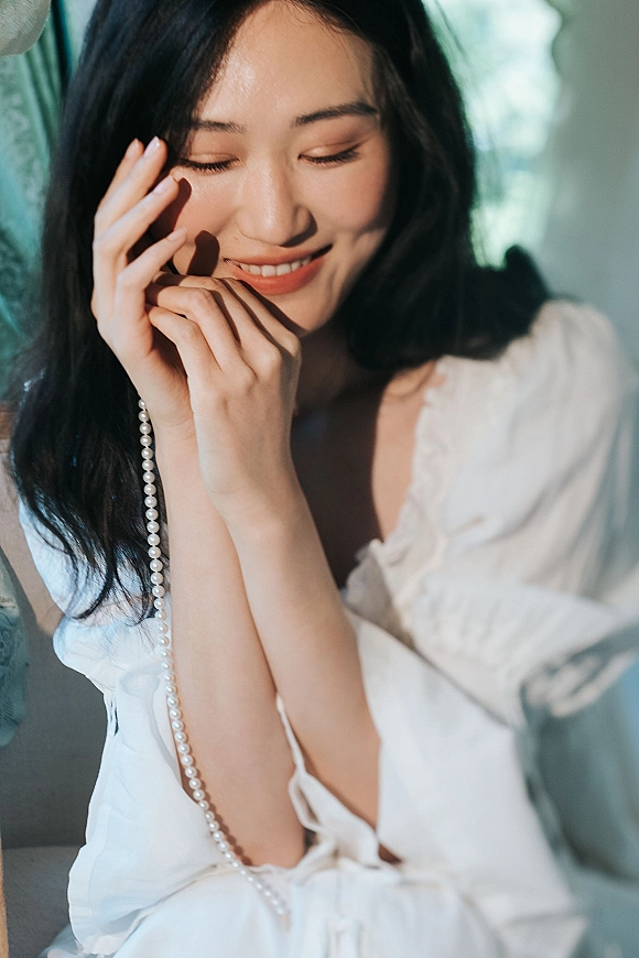 Bridal portrait of a bride getting ready in a white lace robe, hands on face with pearl necklace, softly lit by window light indoors