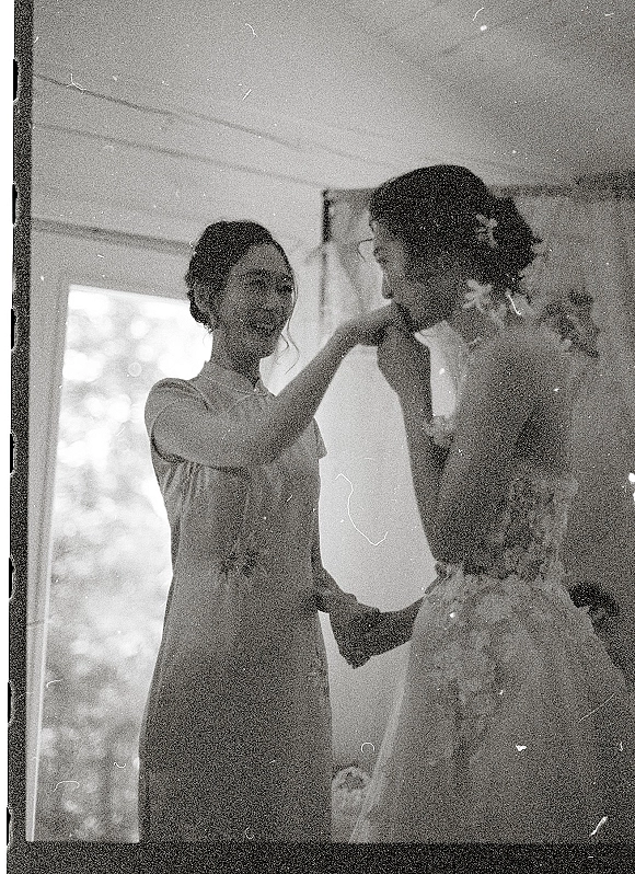 Bride and bridesmaid moment as she gets ready, laughing while the bridesmaid adjusts her bridal veil in a window-lit indoor room
