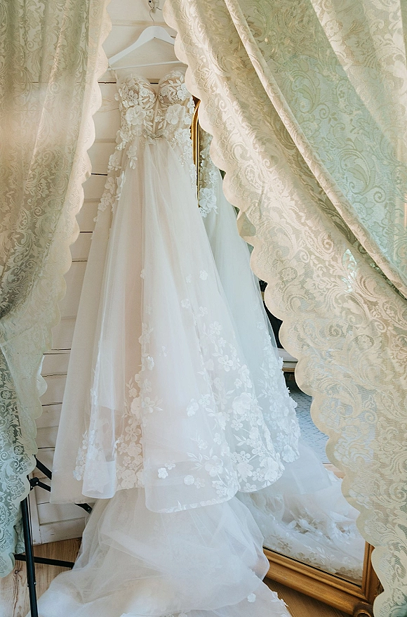 Wedding dress with lace wedding dress details, floral appliqué bodice and tulle skirt on a hanger beside a full-length mirror and curtains