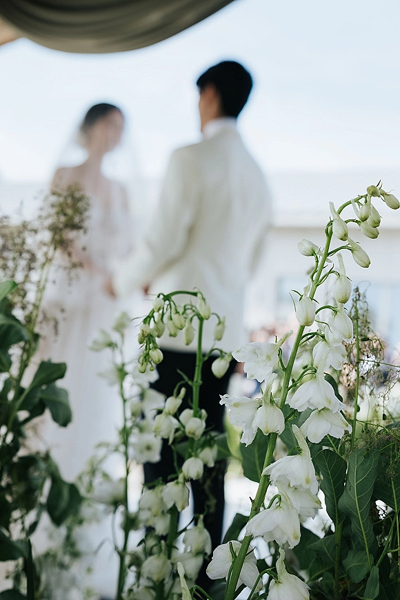 Ceremony moment as bride and groom hold hands under a draped canopy with white florals and greenery on an outdoor terrace backdrop