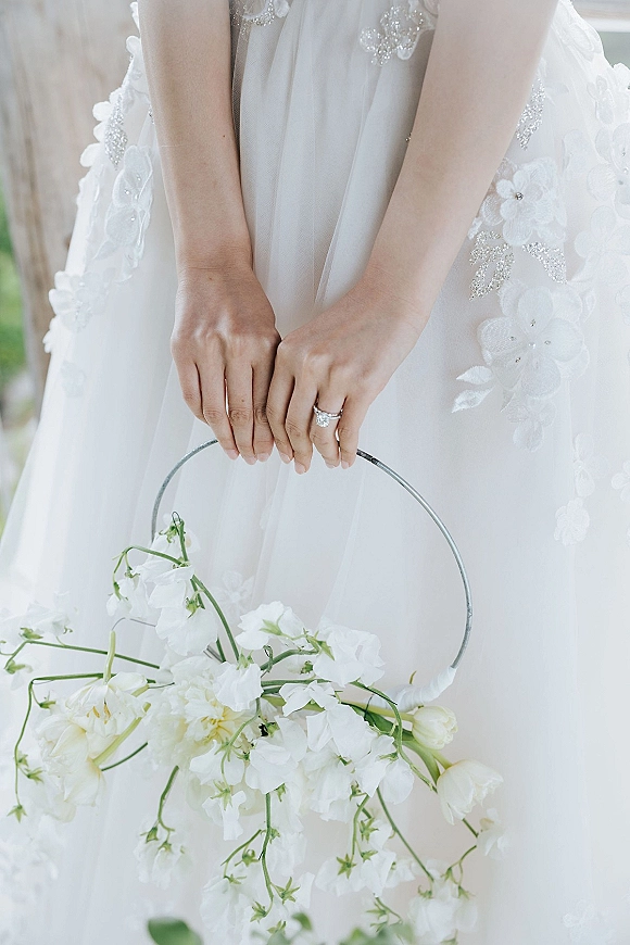 Bridal bouquet of white bridal bouquet blooms held by a bride in a beaded lace wedding dress, with a metal hoop handle and ring visible