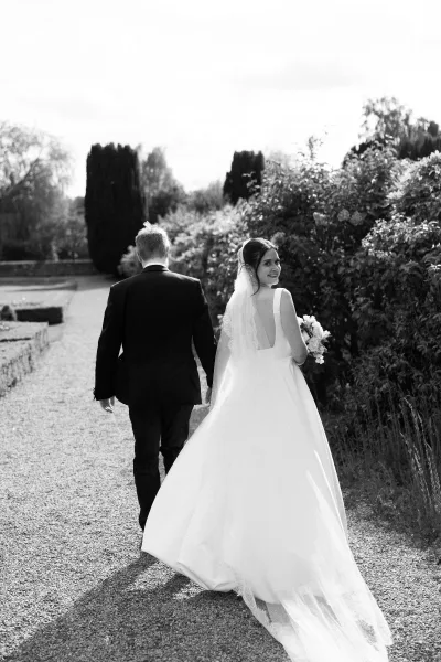Couple portrait of the bride and groom walking along a gravel path, the bride holding a bouquet, framed by garden hedges and trees under open sky.