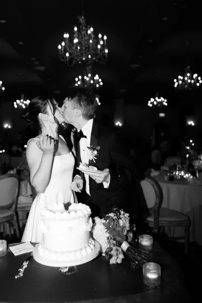 Cake cutting moment as the bride and groom share a wedding cake kiss beside candles in a chandelier-lit reception hall.