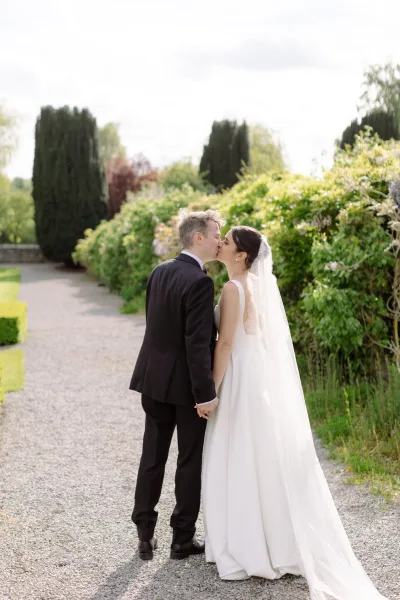 Wedding kiss portrait as the bride and groom kiss, holding hands on a gravel garden path, her veil trailing against manicured hedges.