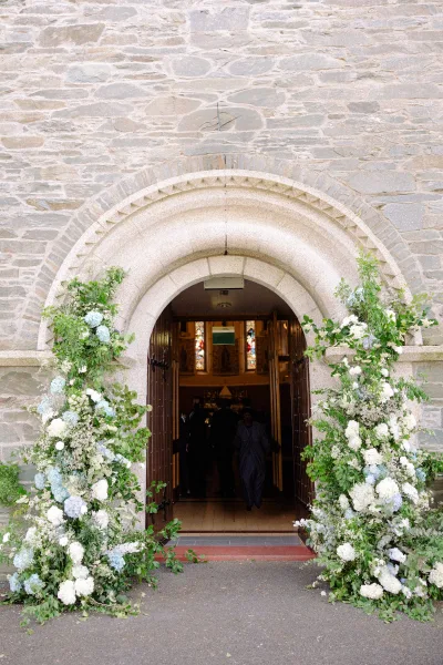 Ceremony entrance flowers frame an arched stone church doorway, lush with blue-and-white hydrangeas beside wooden doors and stained glass.