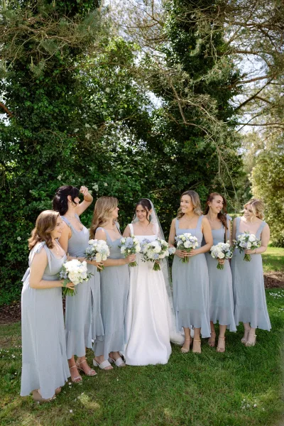 Bride and bridesmaids in dusty blue bridesmaid dresses share a laugh amid garden trees and lush greenery on a sunlit lawn.