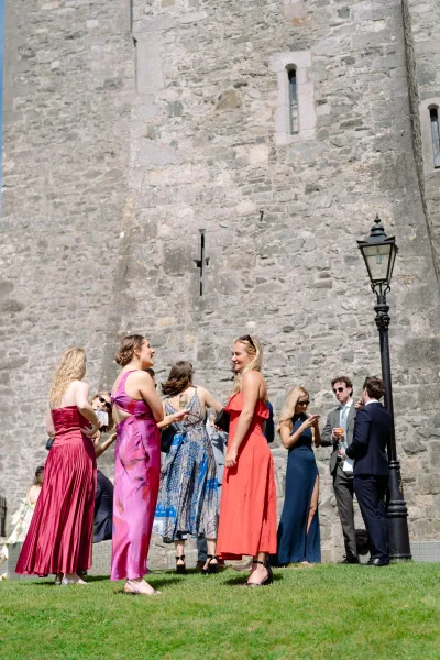 Wedding cocktail hour with wedding guests mingling on a sunlit lawn by a stone wall, toasting with champagne flutes and chatting.