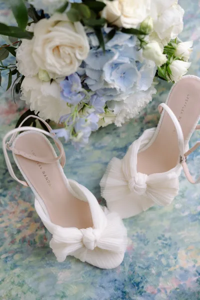 Bridal shoes styled as classic wedding heels set beside a blue hydrangea bouquet on a painterly surface, creating a refined, romantic scene.