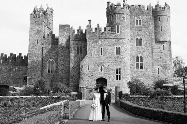 Couple portrait of bride and groom holding hands before a stone castle facade and trimmed garden hedges, the bride cradling a bouquet.