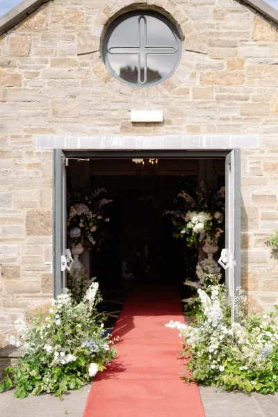 Ceremony entrance decor featuring church doorway flowers in urns, framing an open chapel doorway set in a stone wall.