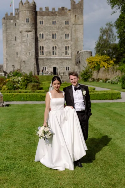 Couple portrait of a bride holding bouquet by a stone castle and garden hedges, the groom in a black tuxedo.