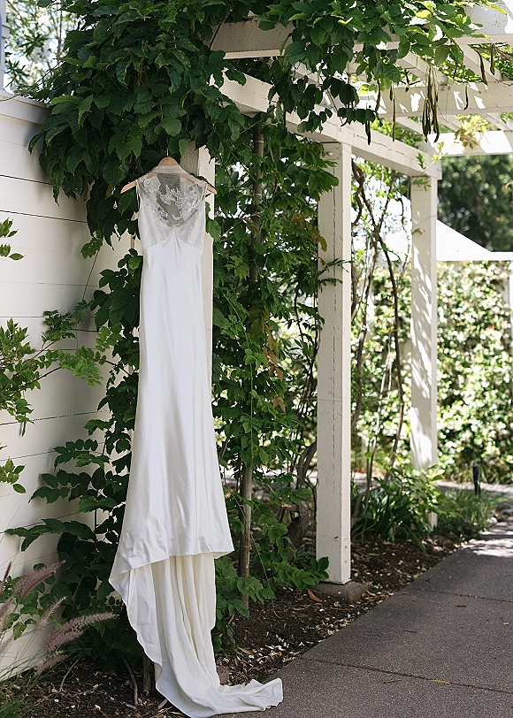 Wedding dress with lace bodice on a wooden hanger, hanging from a white pergola with climbing vines and garden greenery behind