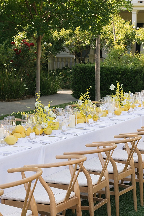 Reception tablescape with white linen place settings, wine glasses, lemons, and yellow-white floral centerpieces on a long garden table