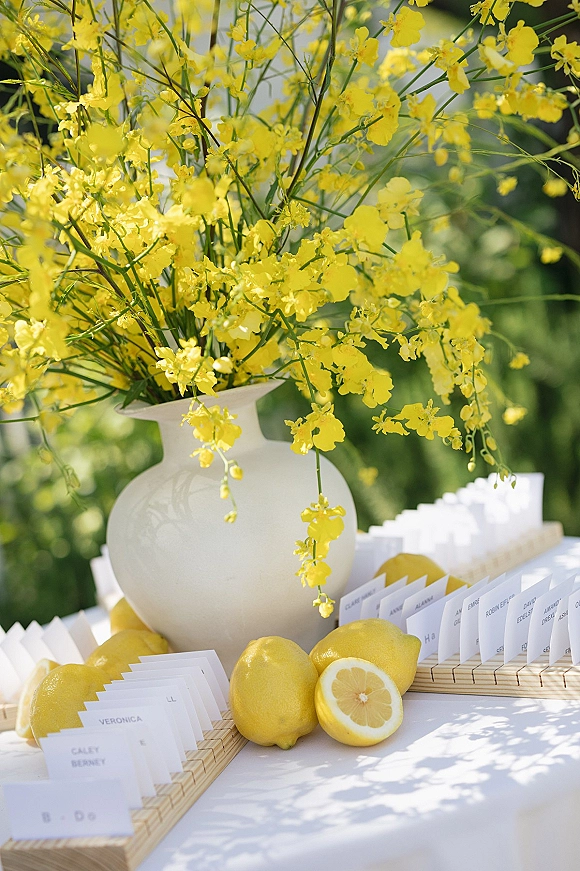 Escort card display with wedding escort cards in wooden holders, yellow flowers in a white vase and lemons on a white table outdoors in sunlight