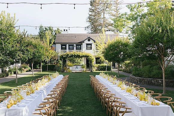 Outdoor reception tablescape with long banquet tables in white linens, wooden chairs, lemon and yellow florals under string lights on a lawn