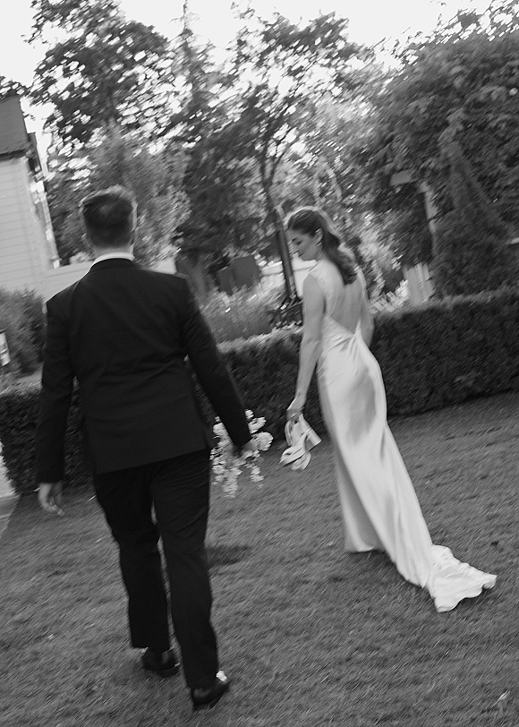 Couple portrait in a black and white wedding photo of bride and groom walking away on a lawn, bride holding heels in hand