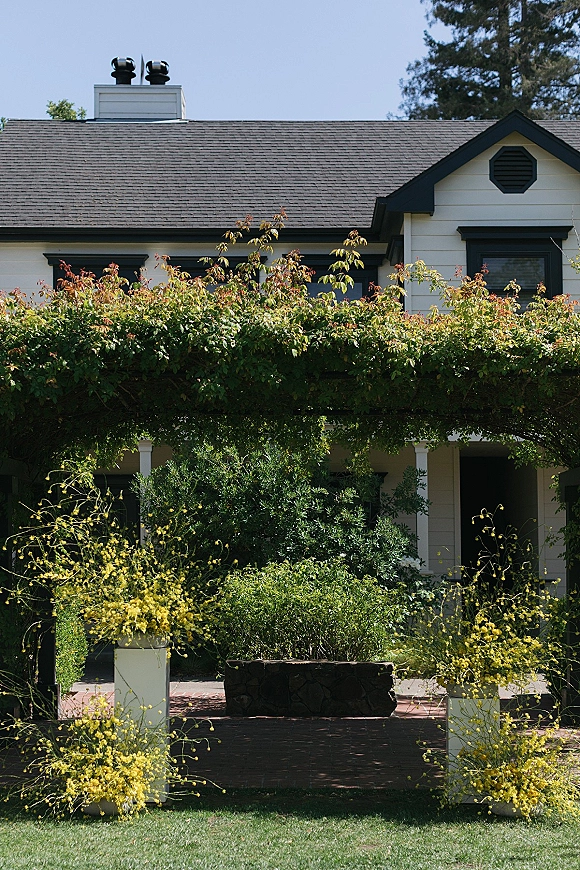 Outdoor wedding ceremony setup with a greenery ceremony arch draped in flowering vines, white pedestals and yellow florals by a porch