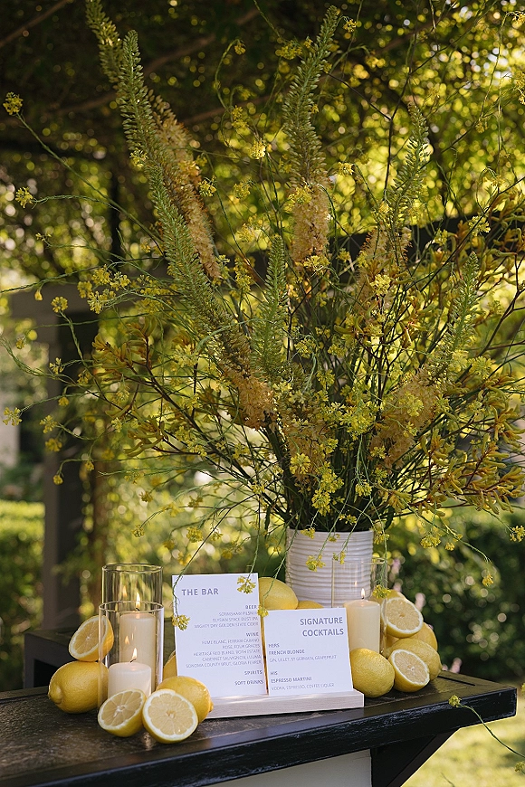 Wedding bar decor with a signature cocktails sign, white menu cards, lemons, florals, and candles against sunlit garden greenery