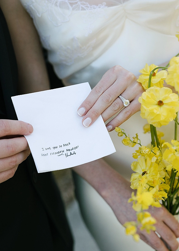 Wedding rings with a diamond engagement ring close up on a bride’s white-manicured hand, beside a handwritten note and yellow flowers on a soft neutral backdrop