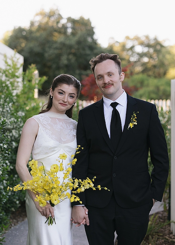 Couple portrait of bride in lace wedding dress holding a yellow flower bouquet beside groom in black suit, in a garden by a white fence