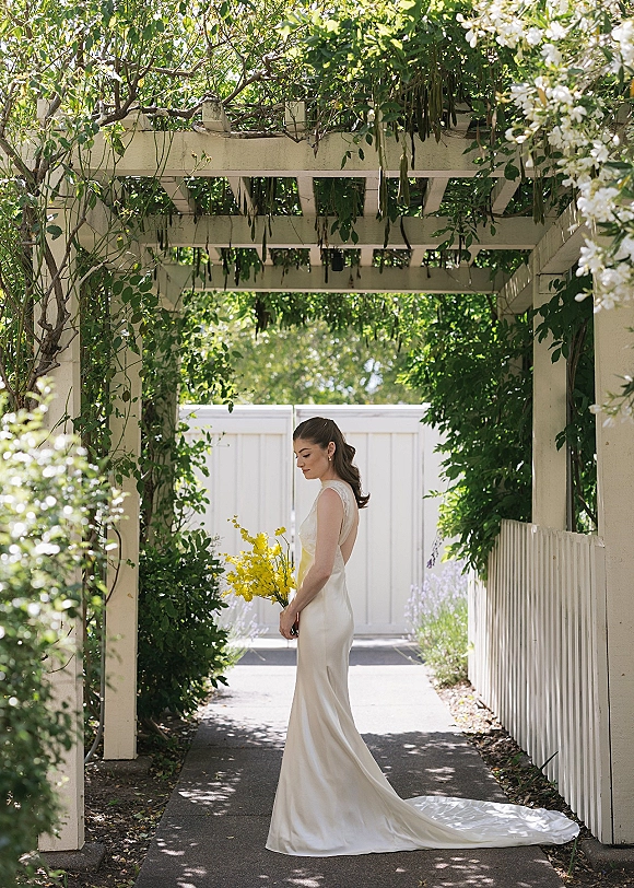 Bridal portrait of a bride holding bouquet in a satin slip wedding dress, side profile on a garden walkway under a vine-covered pergola in dappled sunlight