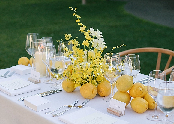 Reception tablescape with lemon wedding tablescape details, yellow and white flowers, lemons, taper candle, and place cards on a green lawn