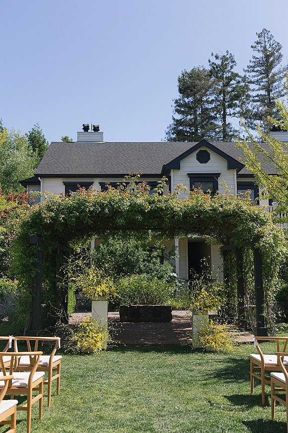 Ceremony setup for an outdoor wedding ceremony with wood chairs lining an aisle runner toward a greenery arch on a lawn by a house patio