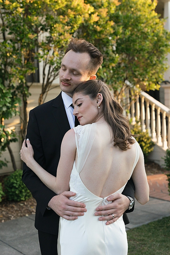Couple portrait of bride and groom embrace, bride in a backless wedding dress looking over her shoulder by a house porch walkway.