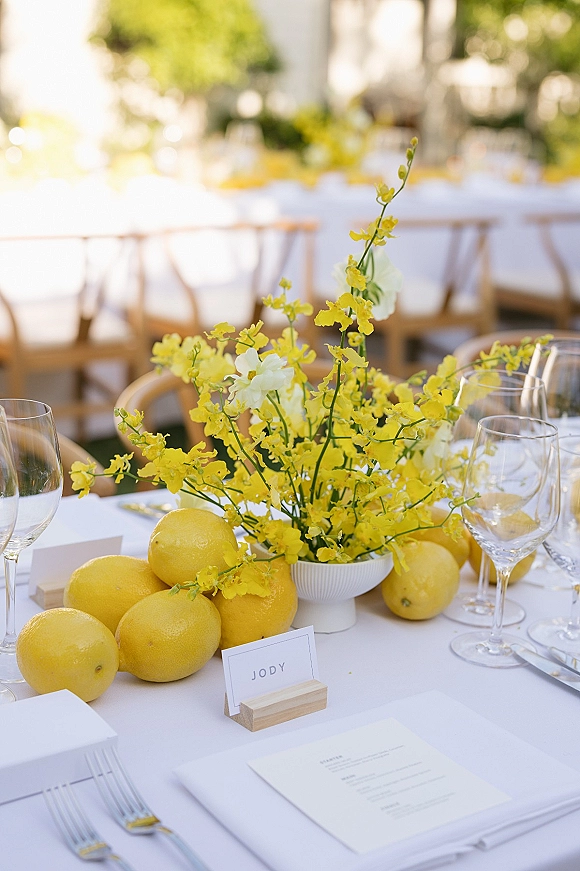 Reception tablescape with a lemon wedding centerpiece of yellow flowers and lemons, white linens and place settings, set outdoors amid greenery