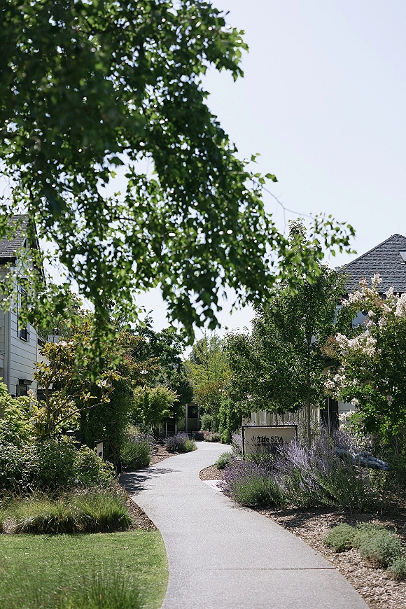 Wedding venue exterior with a venue sign beside a winding walkway, lined with shrubs, flowering bushes, and lavender under blue sky