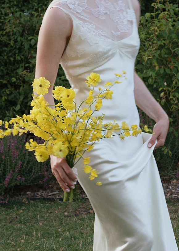 Bridal gown with a lace neckline in satin, paired with a yellow wildflower bouquet, standing on a garden lawn with greenery behind